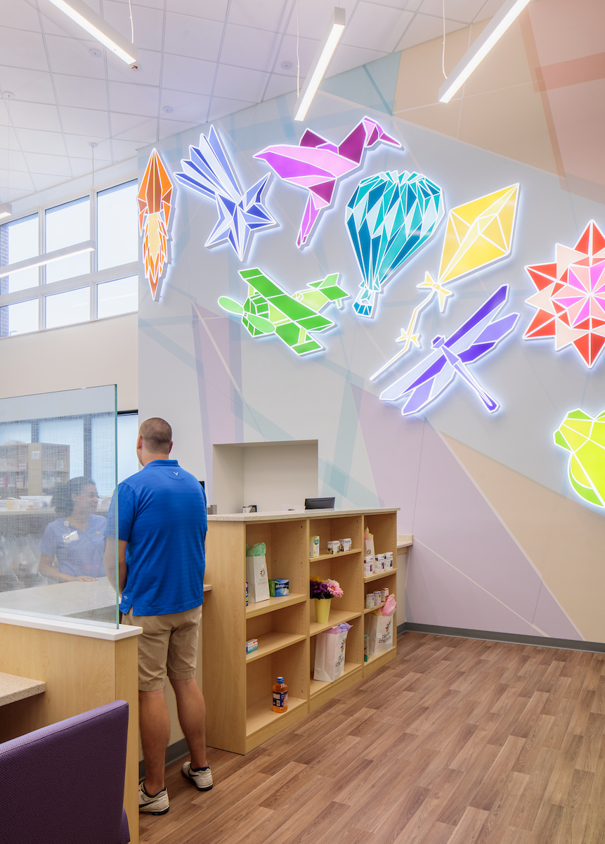 A man in a blue shirt stands at a pharmacy counter with bright, geometric wall art behind him.


