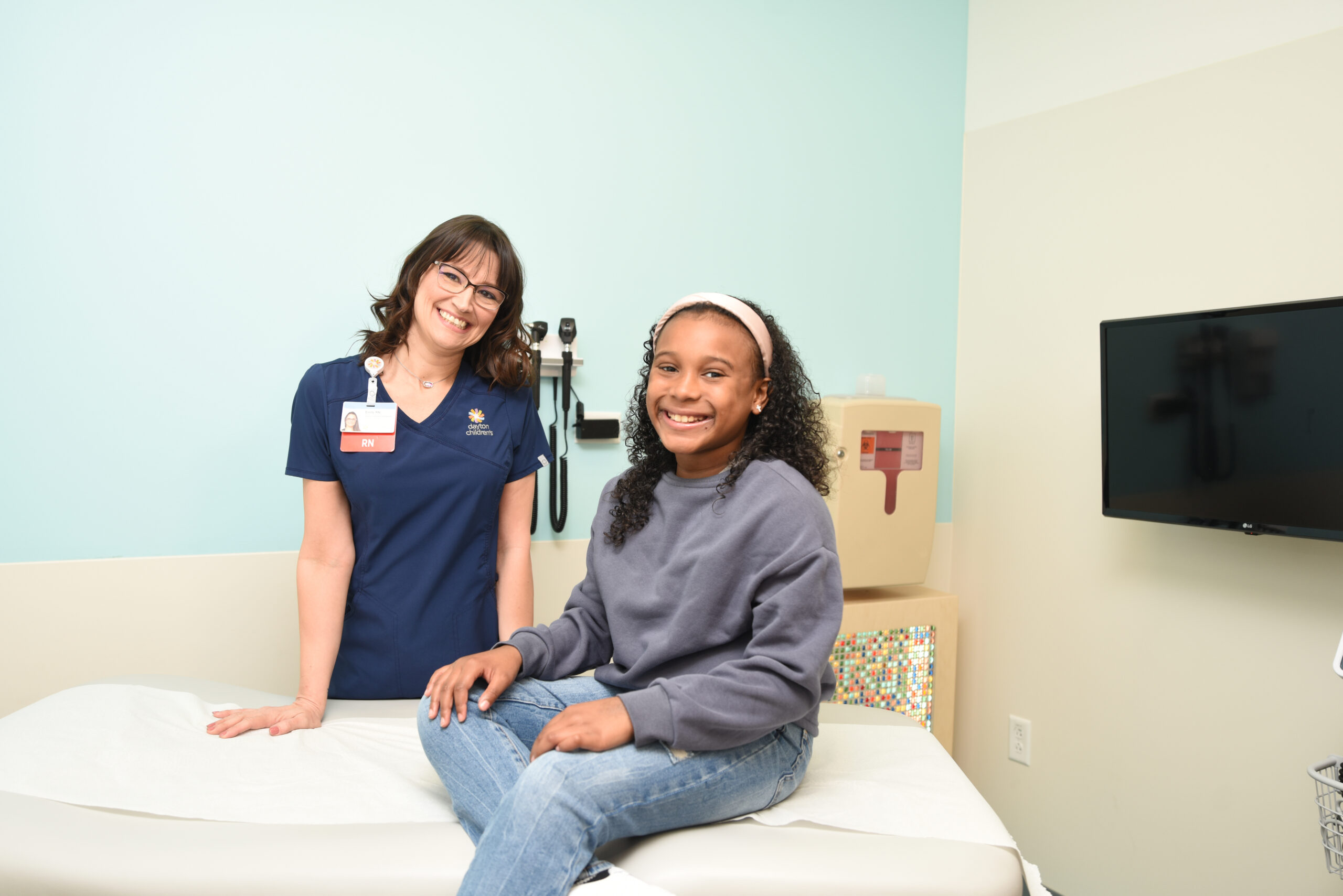 A friendly healthcare provider and a happy young patient smile at the camera while sitting in a modern pediatric exam room
