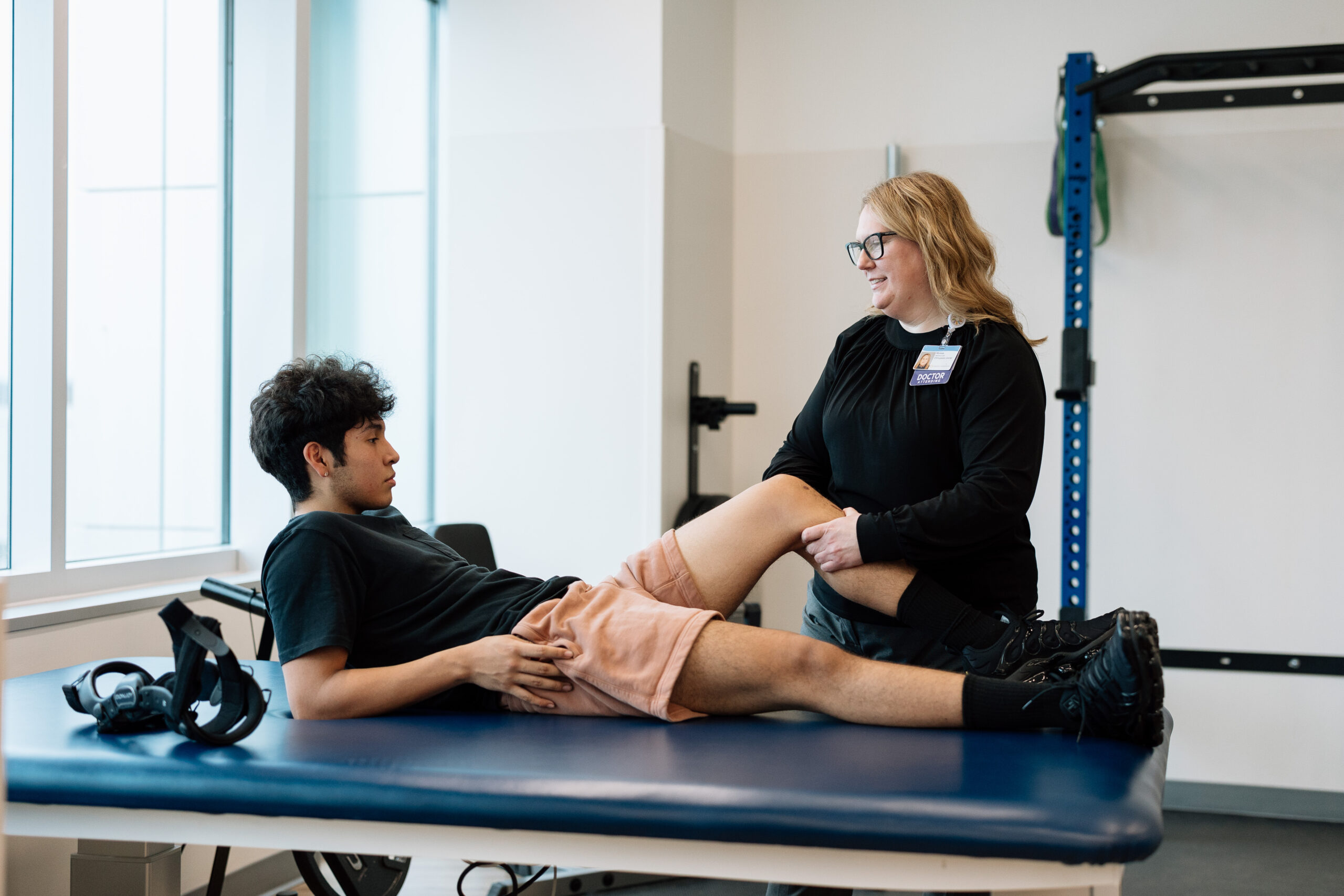 A female therapist helps a male patient with his leg while he lies on a blue physical therapy table.