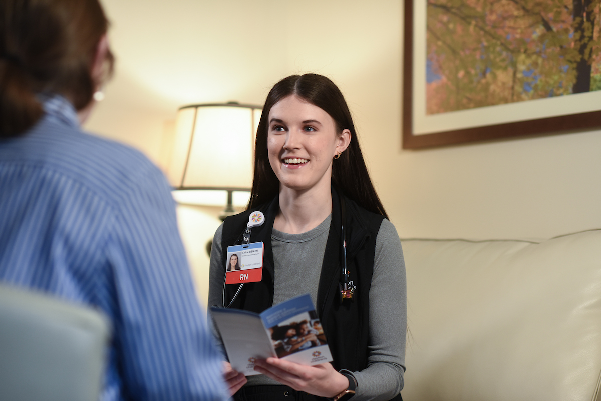 A smiling young female nurse with long dark hair holds a brochure while speaking to someone in a relaxed setting.

