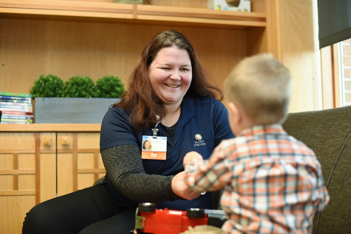 A smiling nurse with a name badge sits in a wooden waiting area, interacting with a young boy in a plaid shirt.

