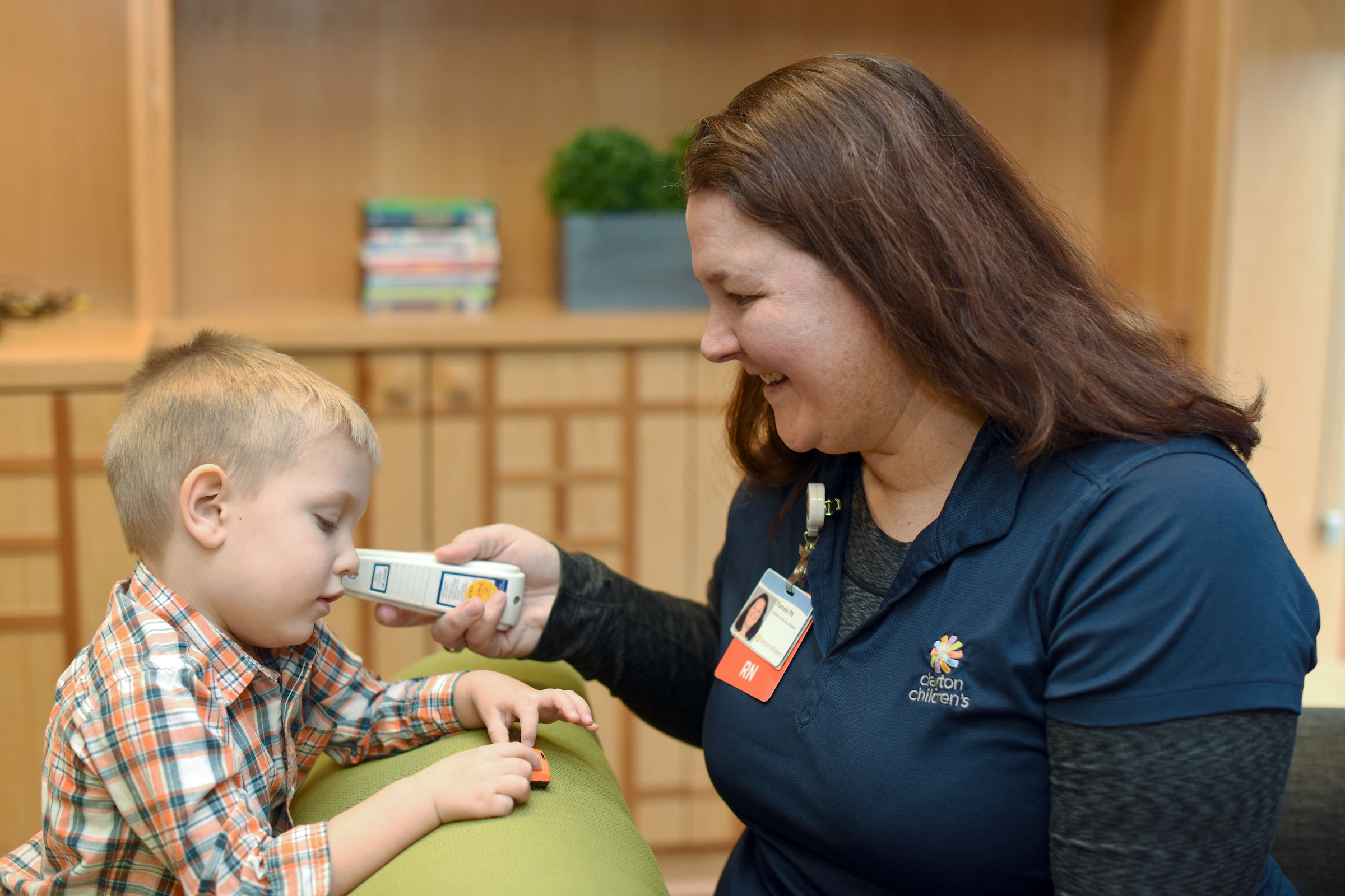 A smiling nurse with a name badge sits in a wooden waiting area, interacting with a young boy in a plaid shirt and holding a hearing device.
