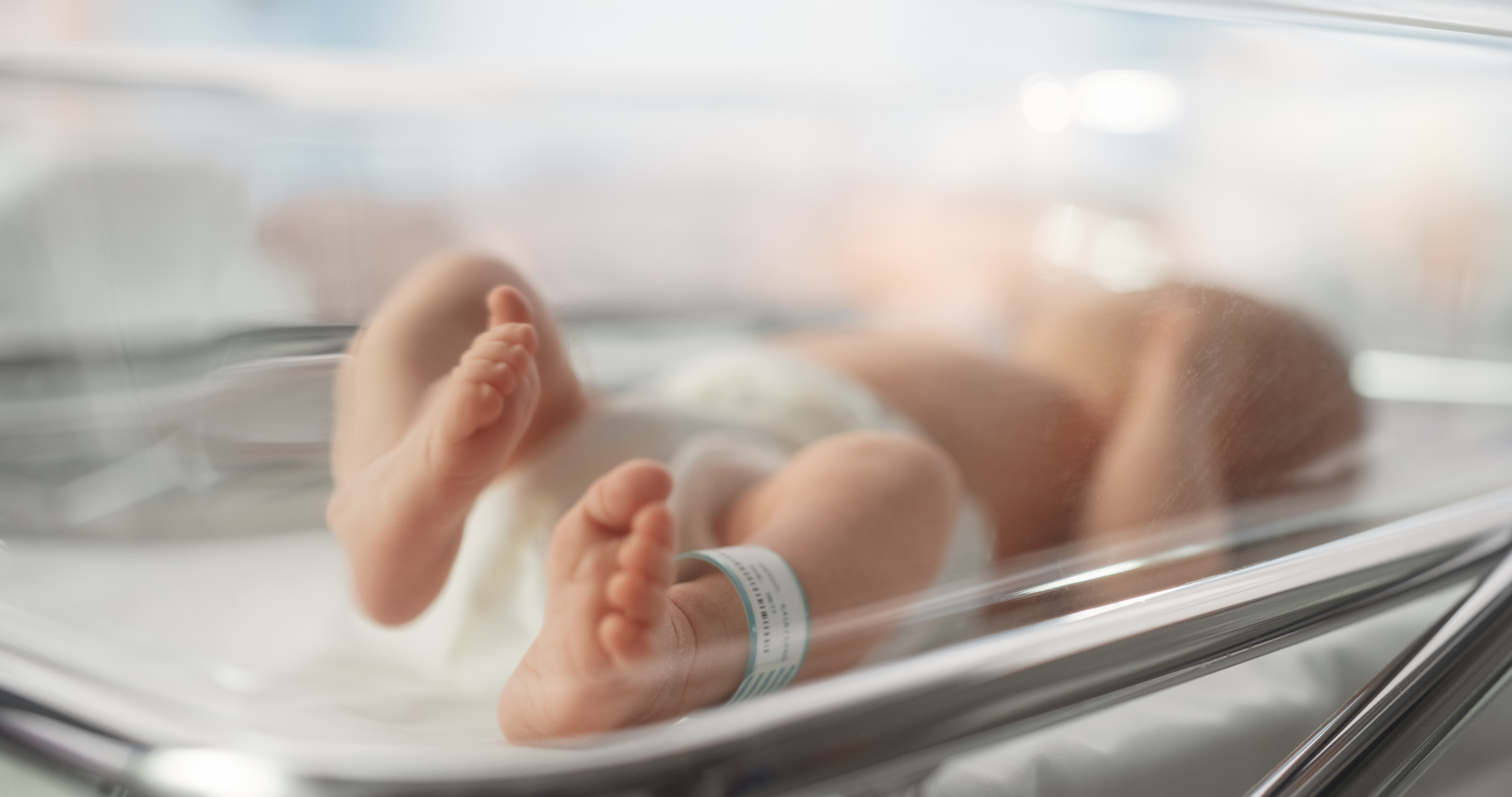 A baby with an identification band on their ankle is lying in a clear hospital bassinet. The baby's feet are in focus, while the rest of the image is slightly blurred.