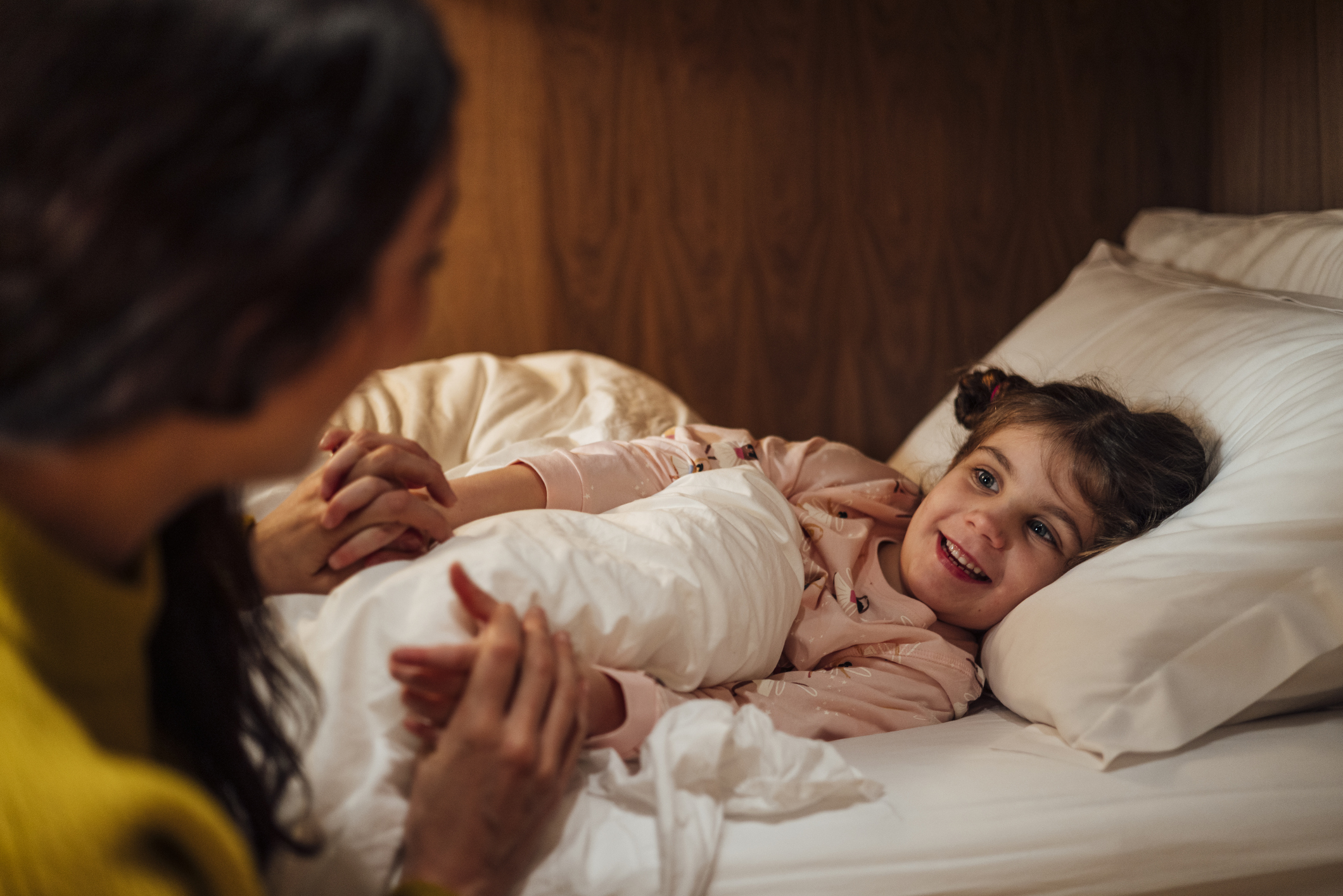 A mother and her young daughter are sitting up in bed, looking at a book together. The girl is holding a teddy bear and laughing with her eyes closed.