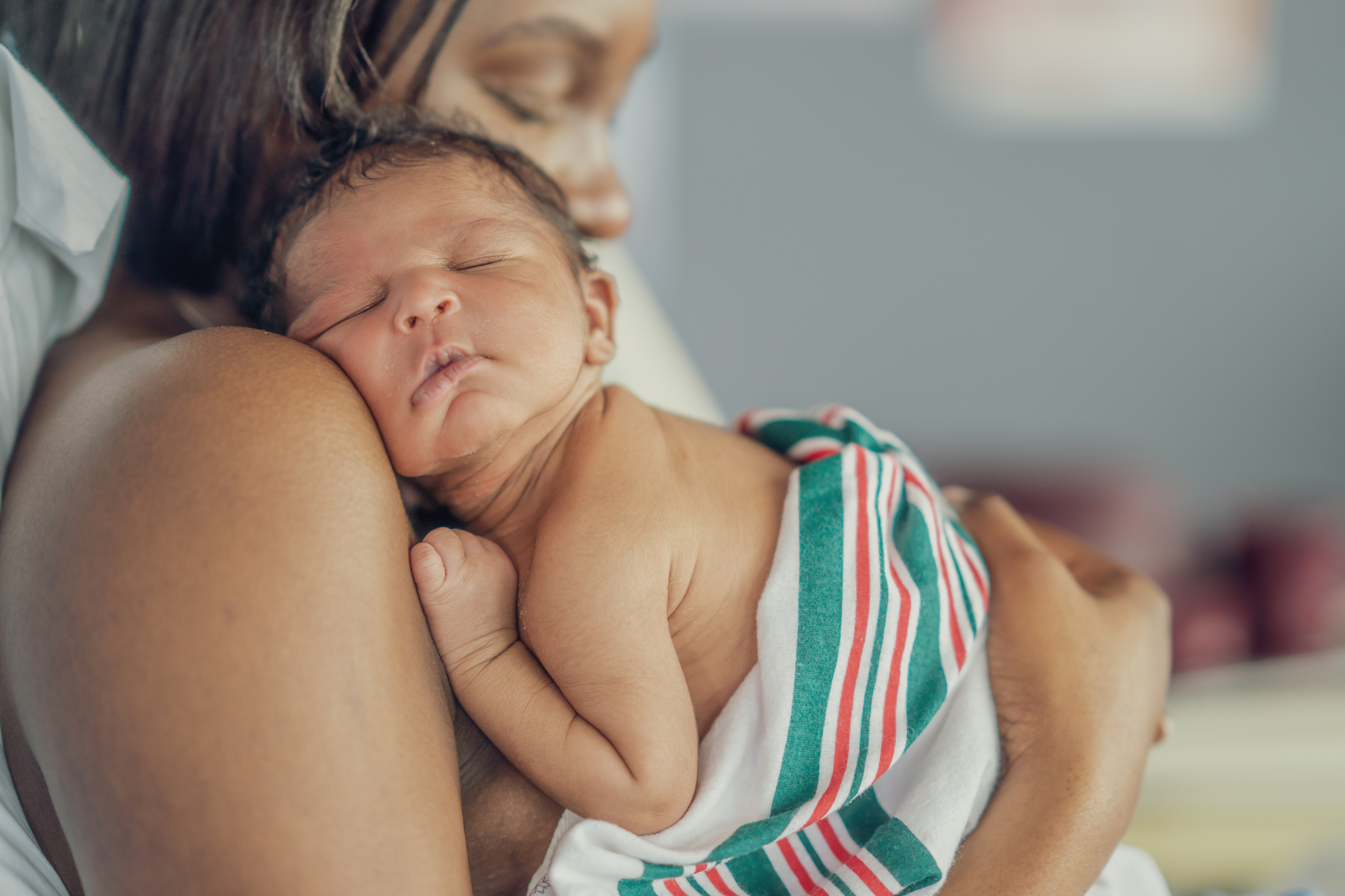 A close-up of a newborn baby with their eyes closed, sleeping peacefully on an adult's shoulder. The baby is partially wrapped in a white blanket with red and green stripes. 

