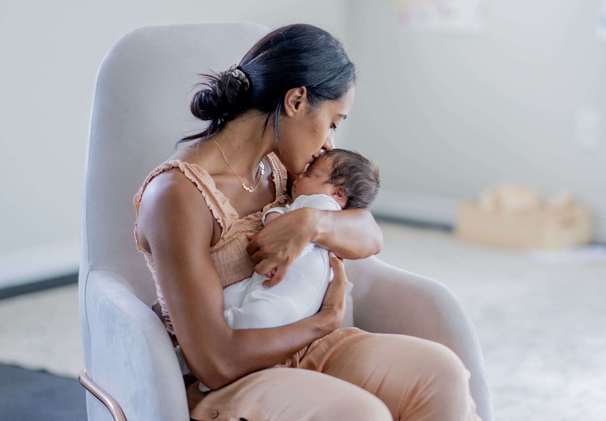 A mother with her hair in a bun sits in a gray rocking chair, holding her newborn baby. The mother is wearing a peach-colored top and pants, and she is kissing the baby's forehead.
