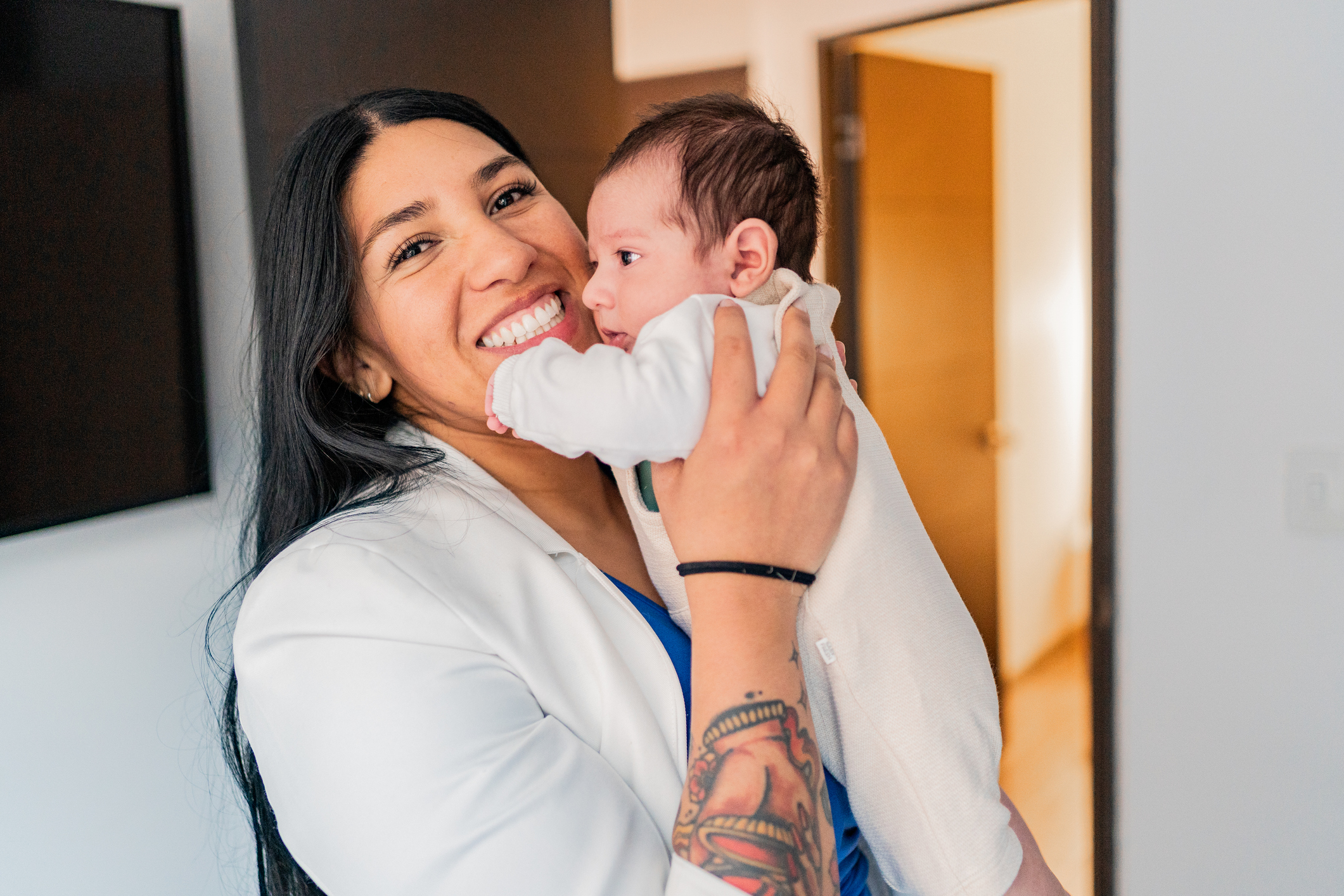 A happy mother with long dark hair smiles at the camera while holding her newborn baby. The baby, dressed in a white onesie, looks off to the side.