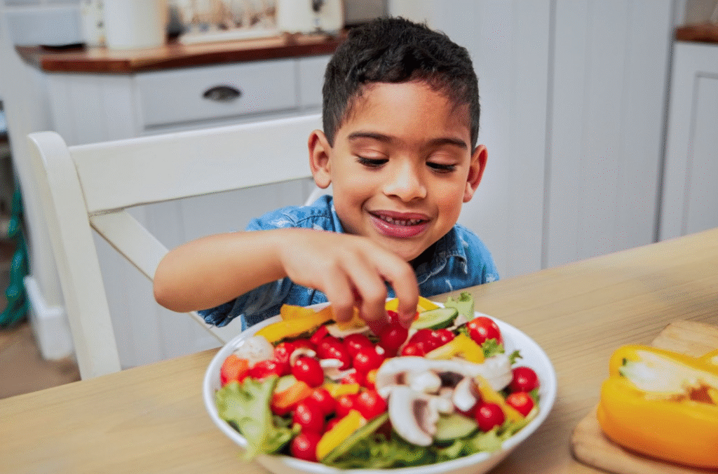 A smiling young boy in the kitchen picks a cherry tomato from a colorful salad in a white bowl.