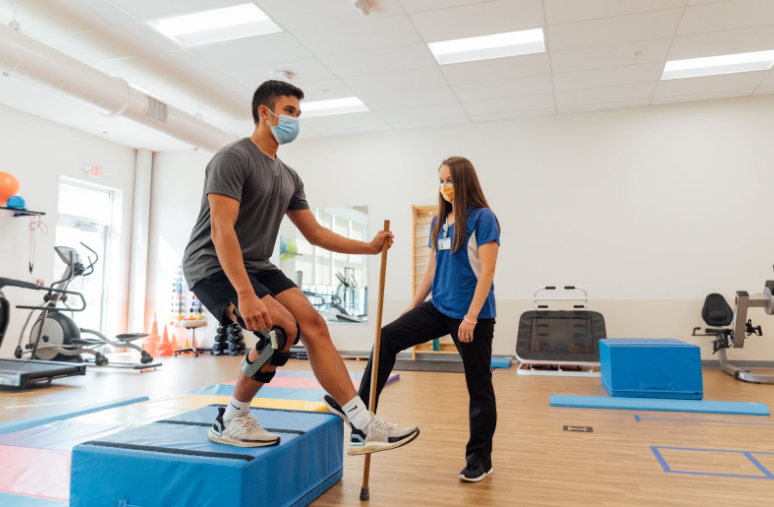 Male athlete wearing a knee brace performing rehab exercises on a step while a female therapist supervises at Dayton Children’s ACL program.