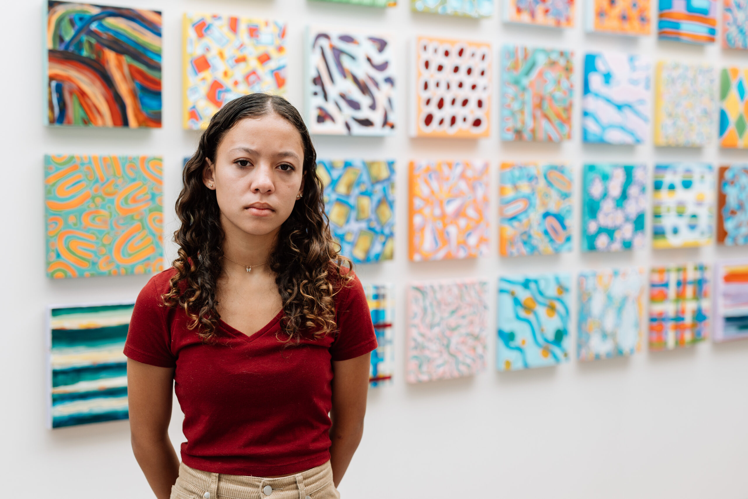 A young woman with curly hair and a serious expression stands in front of a wall adorned with many colorful, abstract paintings.
