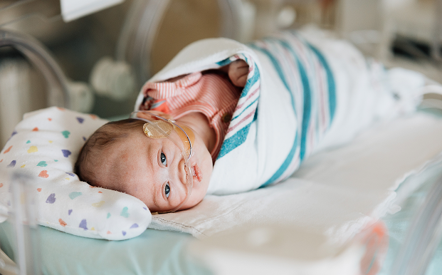 A newborn baby lies swaddled in a hospital crib, symbolizing the impact of donations that support life-saving care for the tiniest patients.