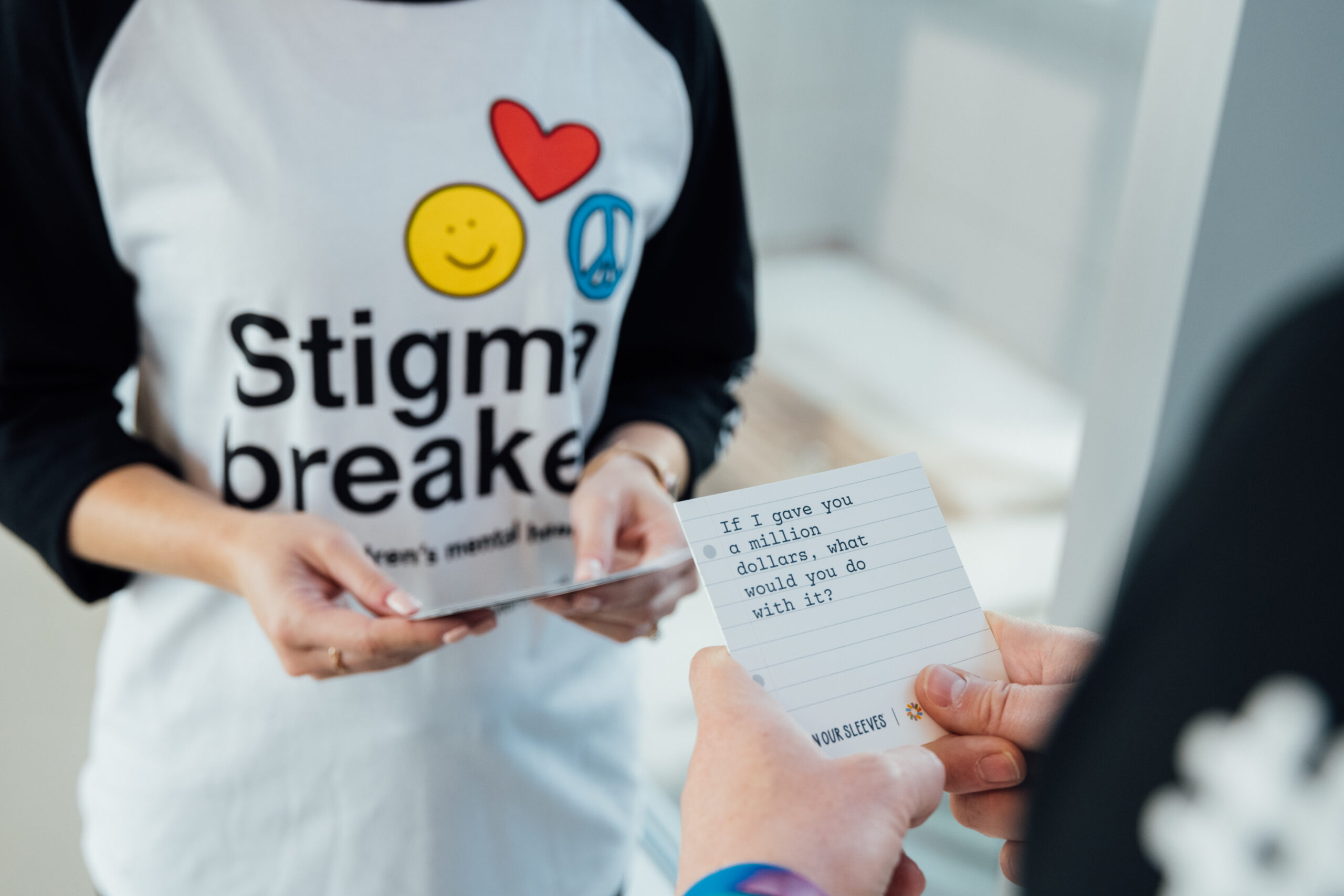 A person wearing a "Stigma breaker" shirt with a heart, smiley face, and peace sign, holds a card that reads "If I gave you a million dollars, what would you do with it?".