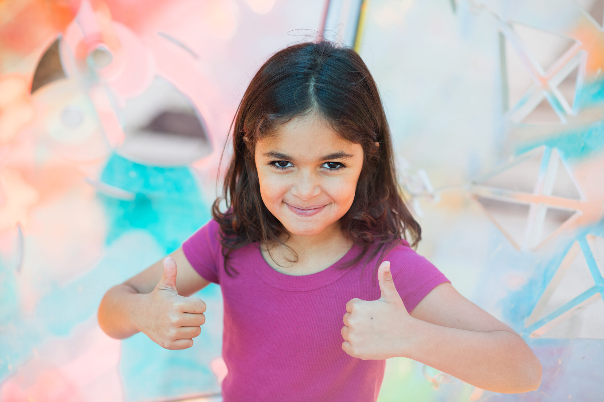 A smiling young girl with long dark hair gives two thumbs up, with a blurry, colorful background.