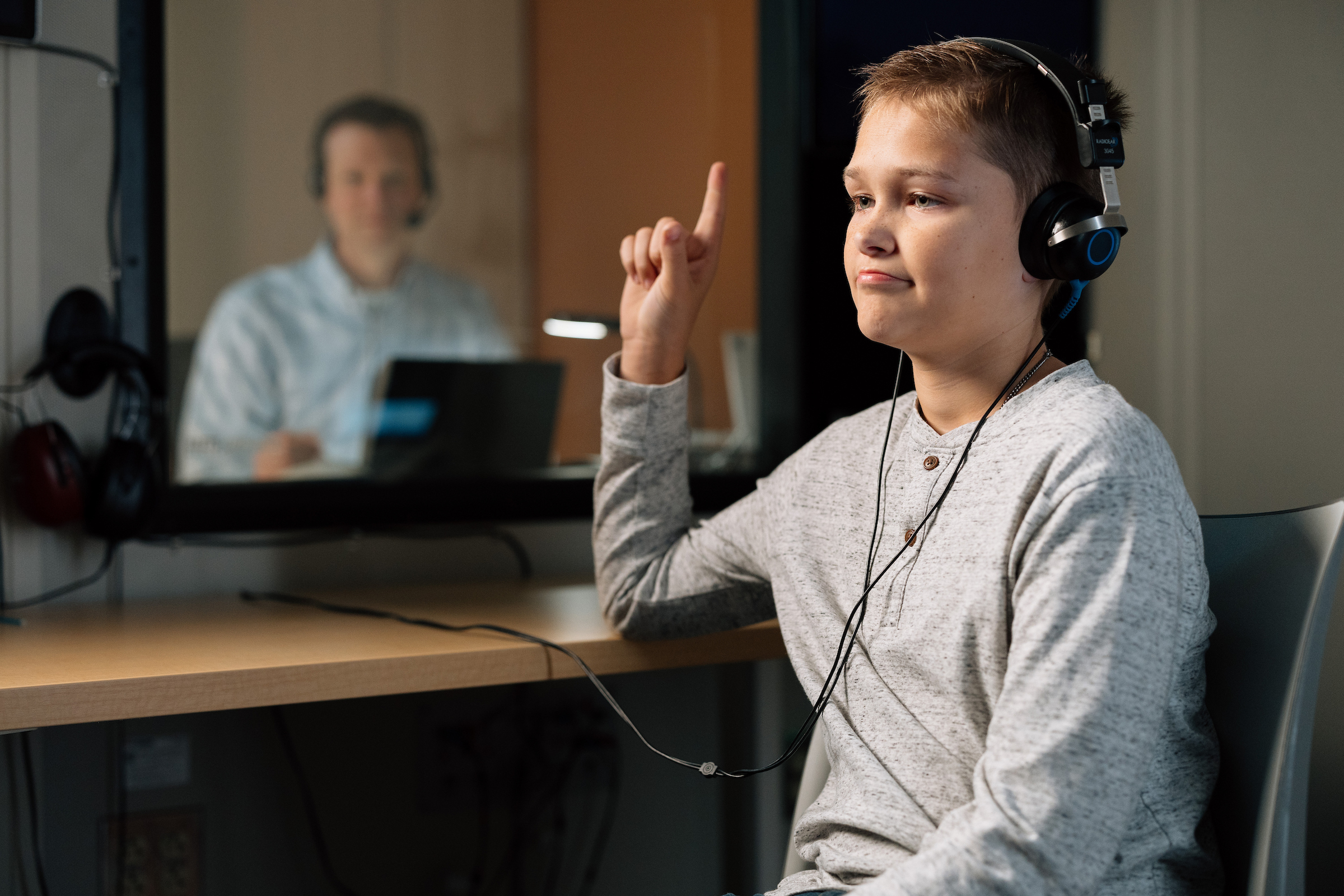 A boy wearing headphones raises his finger, indicating he hears something, during an audiology test.
