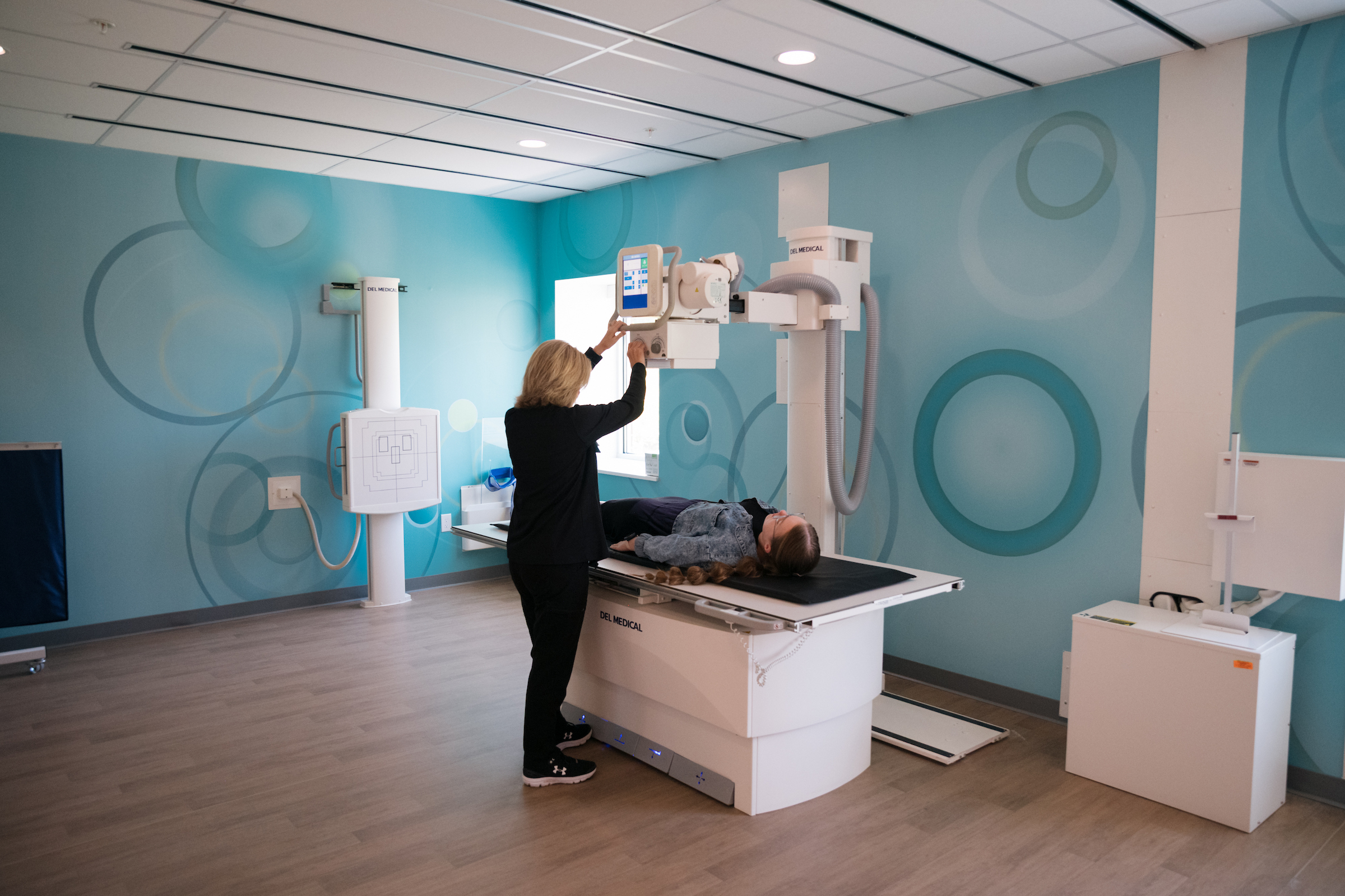 A medical professional positions an X-ray machine over a patient lying on a table in a room with blue patterned walls.