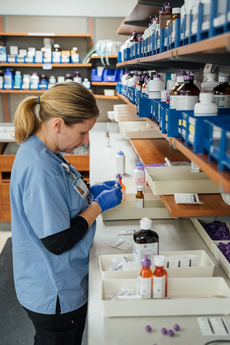 A female pharmacist in blue scrubs and gloves carefully prepares medication bottles in a well-stocked pharmacy.

