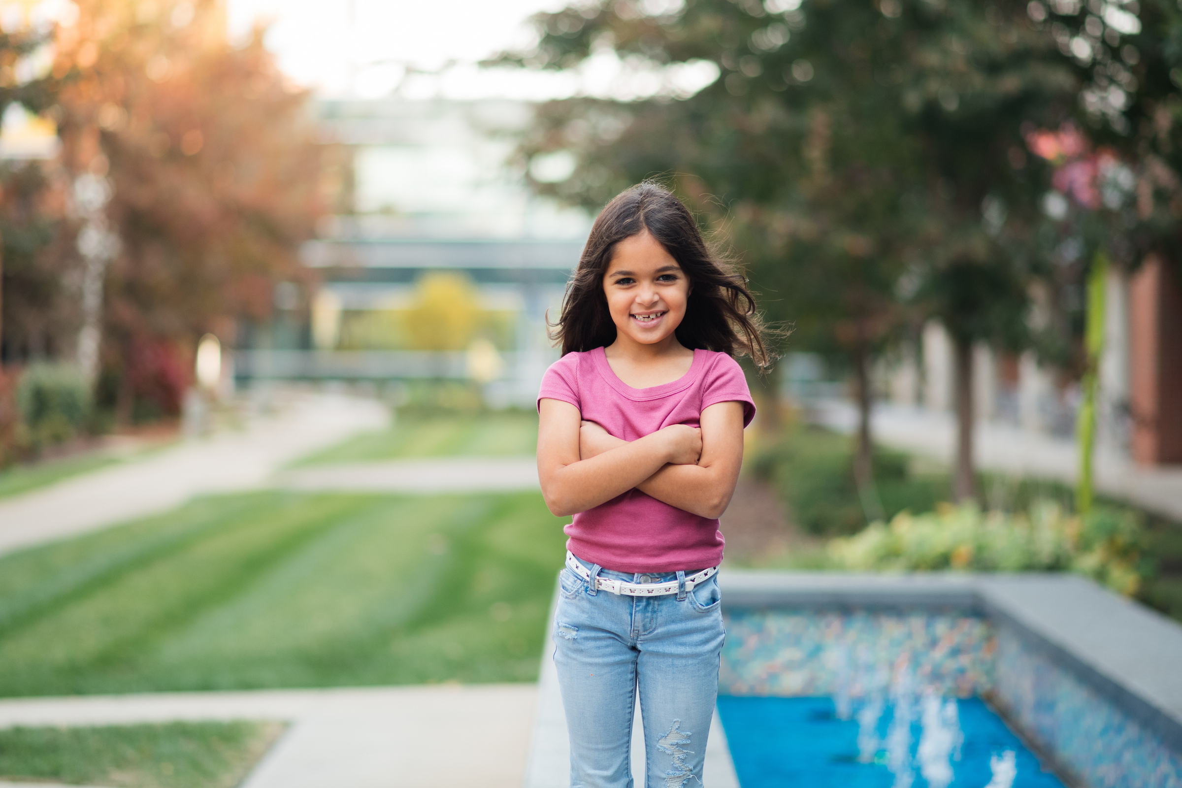 A happy young girl with long dark hair, wearing a pink t-shirt and jeans, stands with crossed arms in a park-like setting.