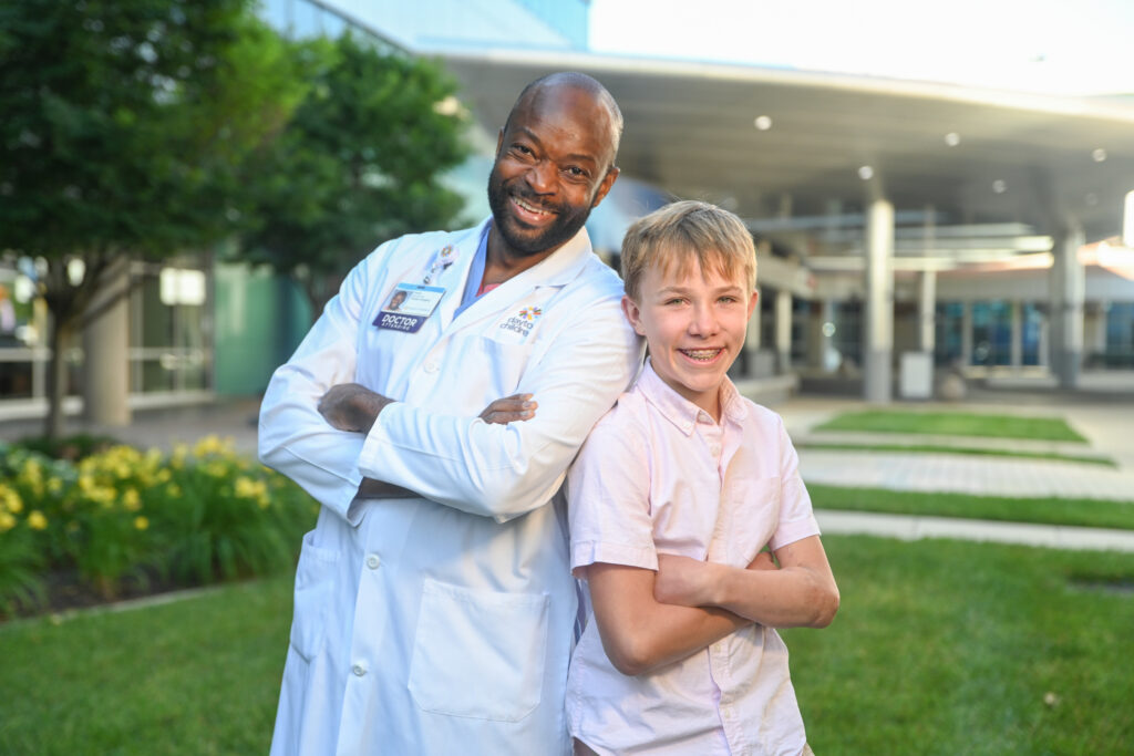 A grinning doctor in a lab coat and a smiling boy with braces, both with crossed arms, lean back-to-back outdoors.

