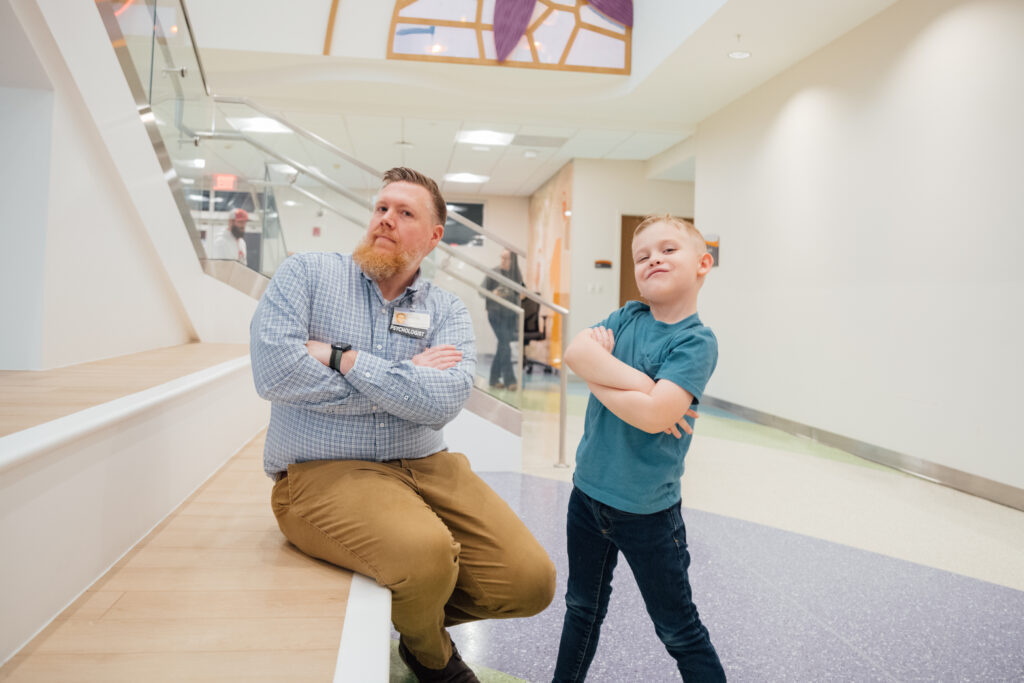 A male pediatric psychologist takes a confident and happy pose with a young boy who is smiling. 