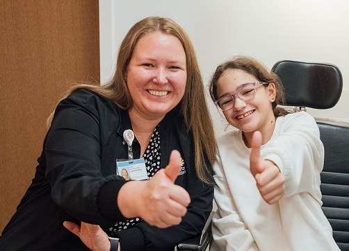 A smiling male doctor in a white lab coat sits while a young girl with pigtails stands behind him, in a bright, modern room.