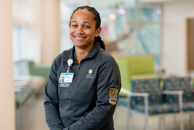A smiling healthcare worker stands in a bright hospital setting, representing career opportunities and the welcoming environment at a pediatric care facility.