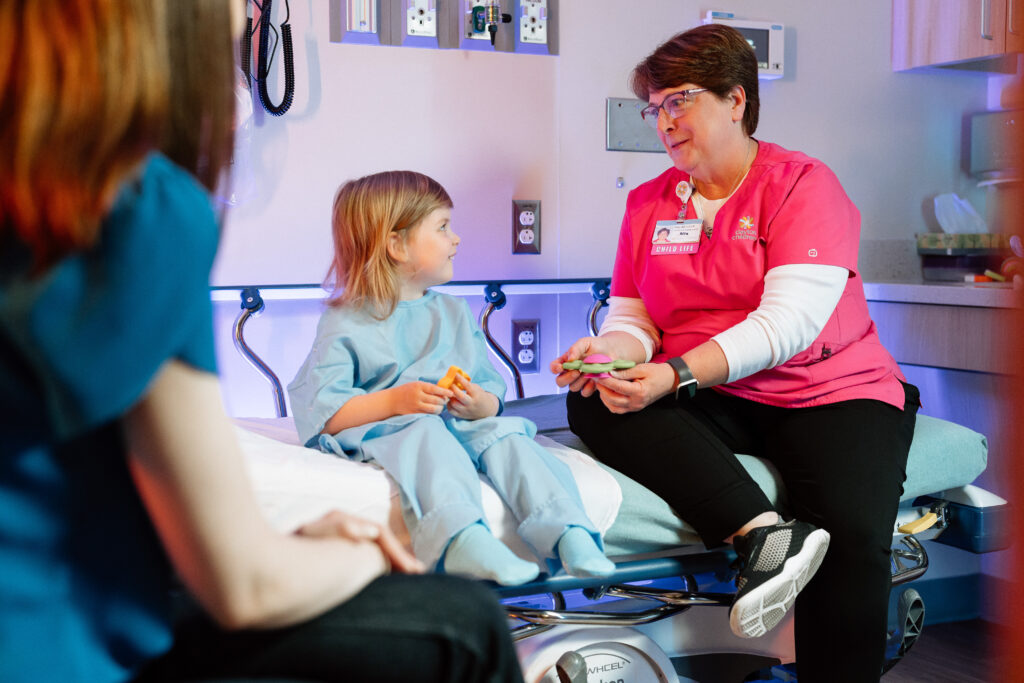 A child life specialist in pink scrubs talks to a young girl in a hospital gown, holding toys, while a third person is partially visible.