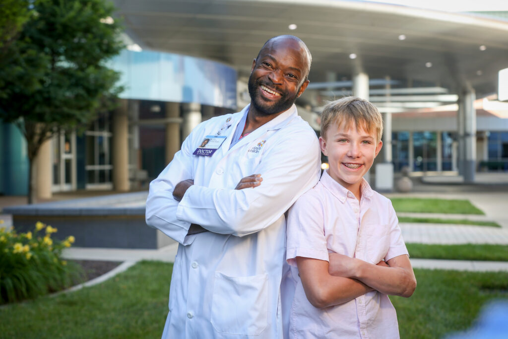 A grinning doctor and a smiling boy with braces, both with crossed arms, lean back-to-back outdoors.