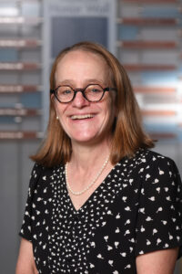 A professional headshot of a smiling woman with shoulder-length brown hair, wearing tortoiseshell glasses, a pearl necklace, and a black and white patterned top. 