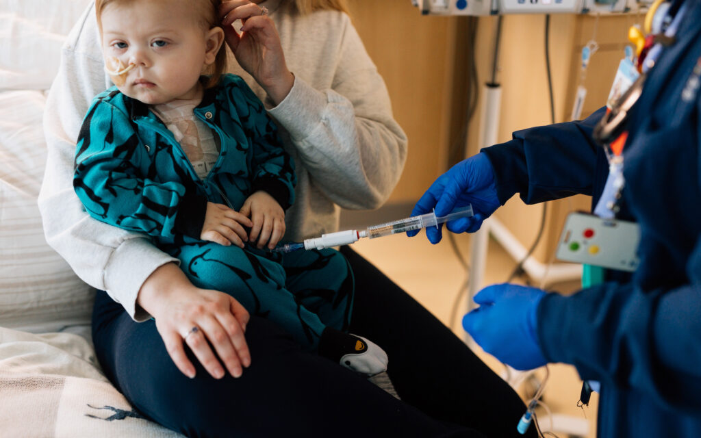 A young child with a nasal tube sits on a caregiver’s lap while a nurse in blue gloves administers medication through a syringe.
