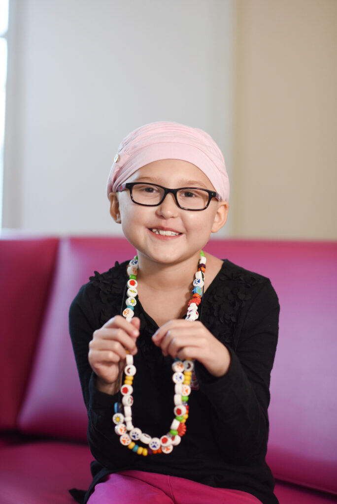 A young girl wearing glasses and a pink headscarf smiles while holding a colorful bead necklace at Dayton Children’s.
