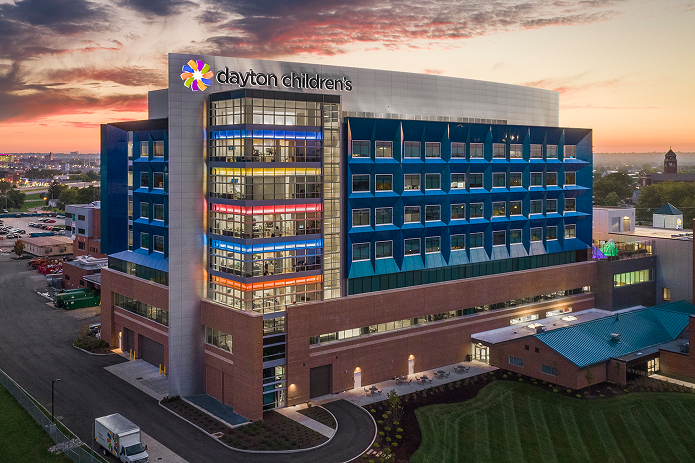 An impressive modern hospital building, "dayton children's," glows with colorful lights against a dramatic sunset sky.