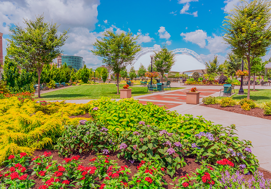 A vibrant park scene with colorful flower beds, green lawns, trees, pathways, and a large white arched structure in the background under a blue sky with clouds.
