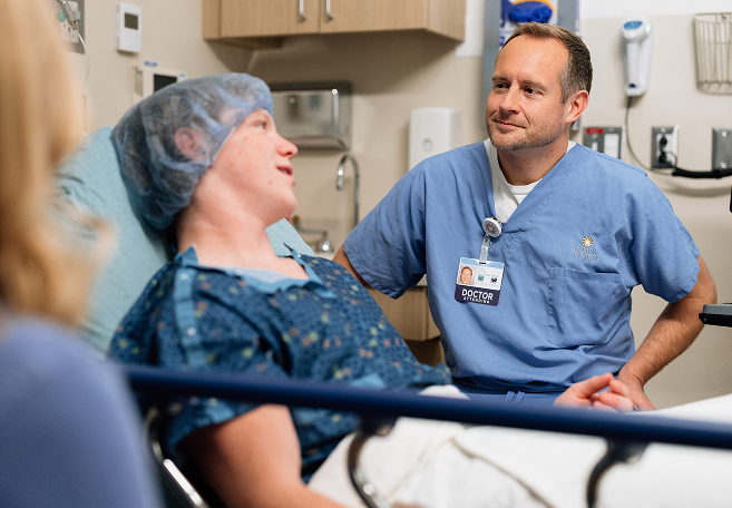 A male doctor in blue scrubs and a patient in a hospital gown and hairnet are smiling and looking at each other while a third person's shoulder is visible in the foreground.
