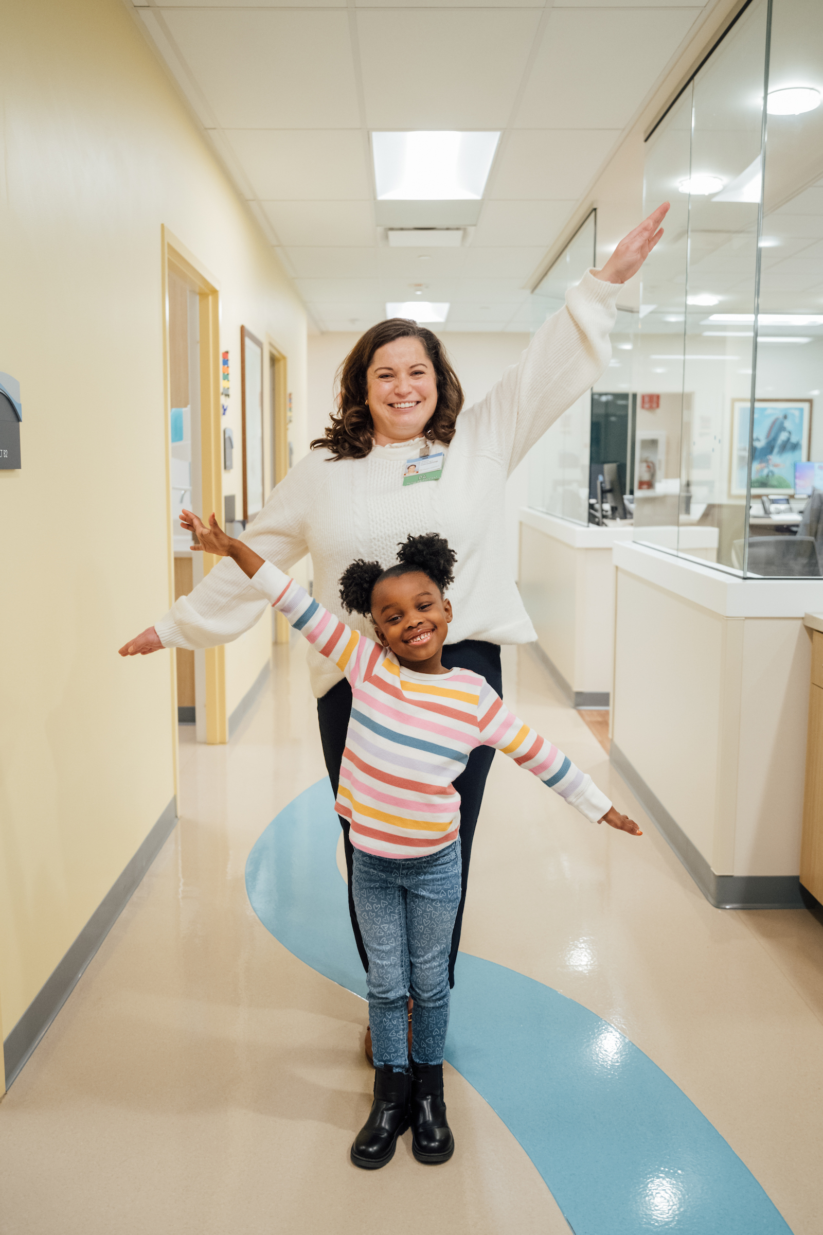 Child and caregiver joyfully walking down a hospital hallway, arms outstretched and smiling.