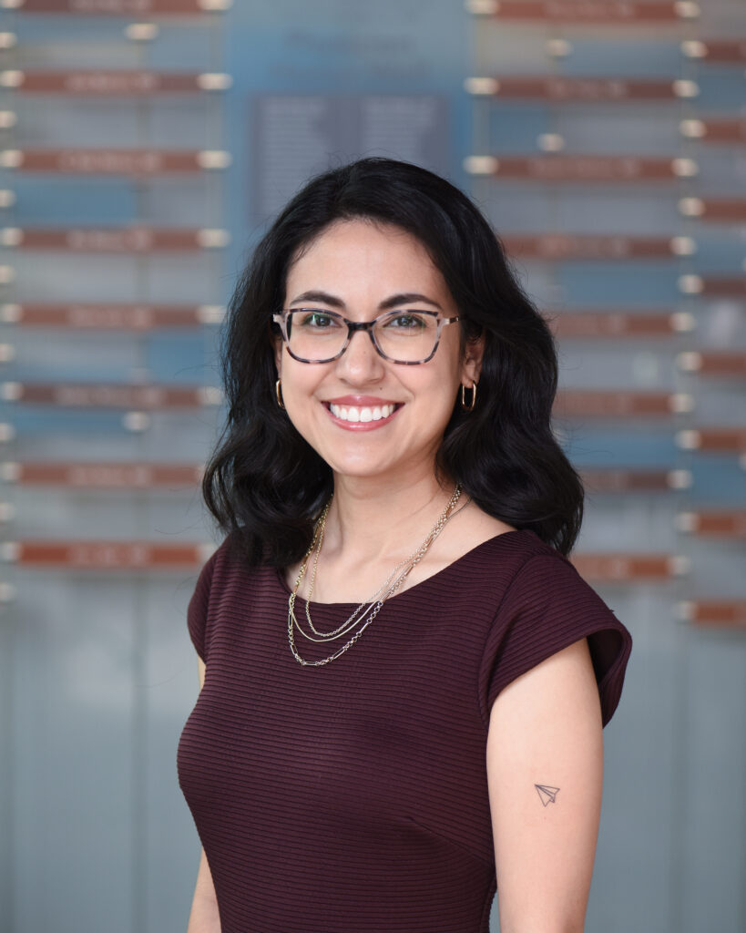 A headshot of a smiling woman with dark, wavy hair, wearing cat-eye glasses and a burgundy top, with a paper airplane tattoo on her arm.