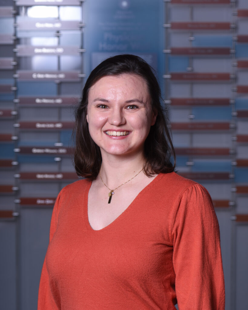 A friendly professional headshot of a smiling woman with medium-length dark hair, wearing an orange V-neck sweater and a delicate necklace.
