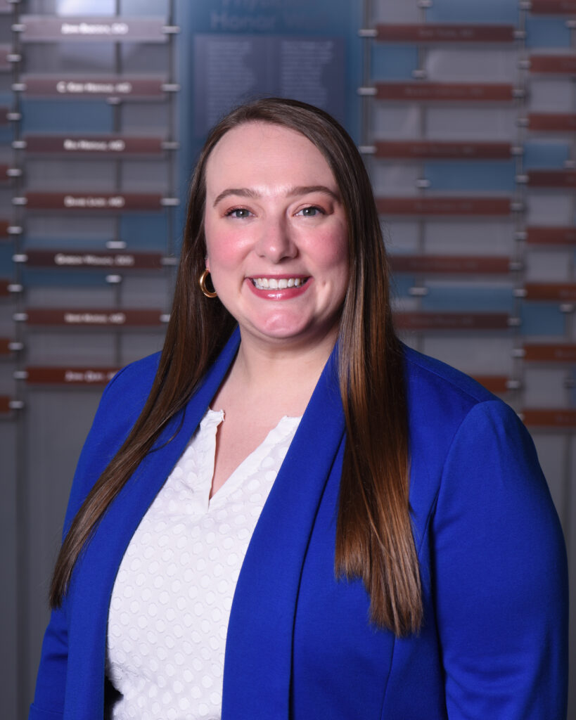 A smiling woman with long brown hair wears a bright blue blazer over a white patterned top in a professional headshot.