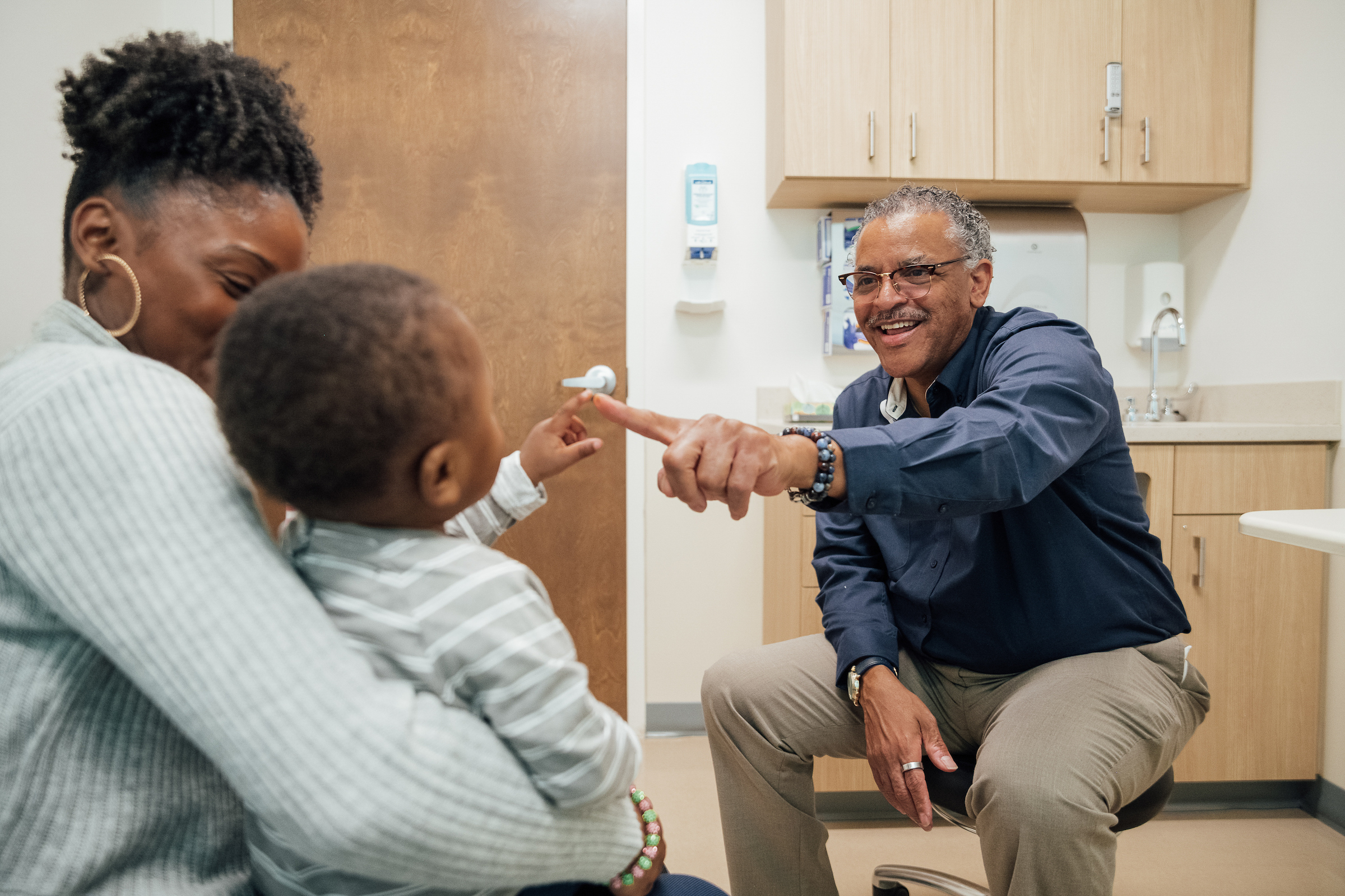 Smiling doctor interacting with a young child sitting on a parent’s lap during a medical visit.