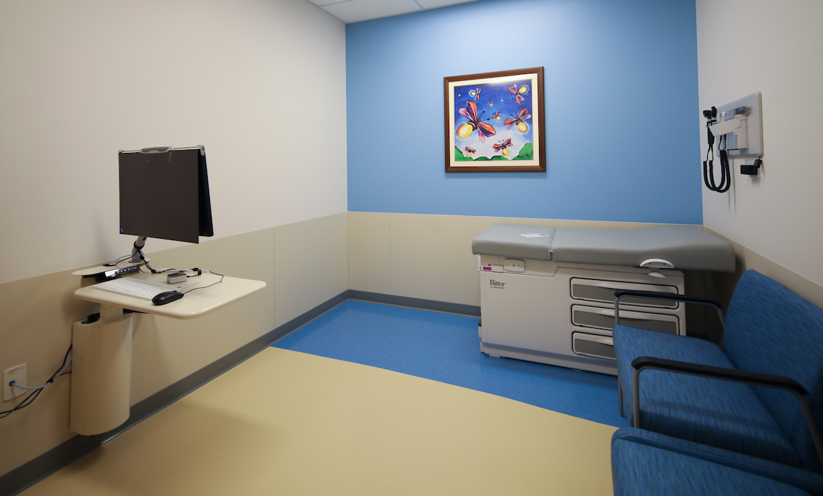  Bright and clean pediatric exam room with a medical table, computer workstation, blue chairs, and colorful butterfly artwork on the wall.