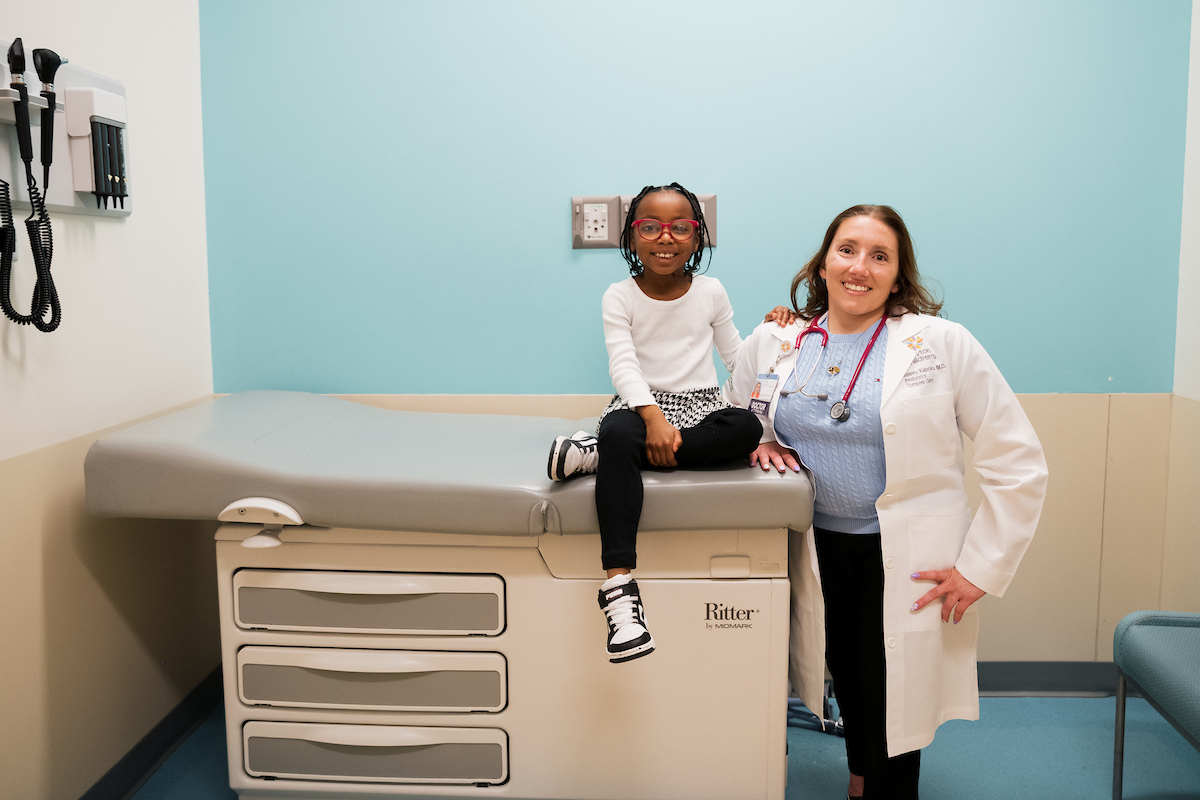 Smiling pediatrician standing beside a young girl sitting on an exam table in a brightly colored medical room.