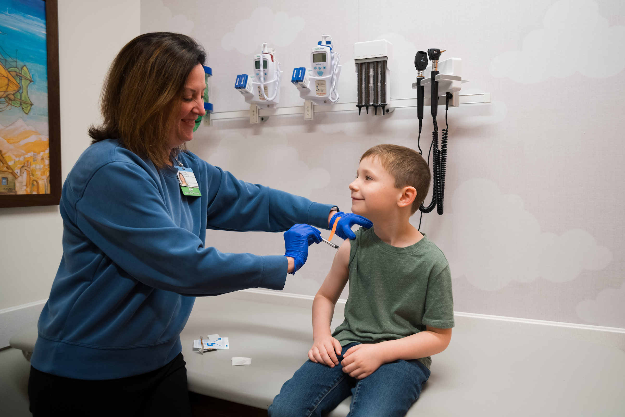 Smiling nurse gives a young boy a vaccination in a bright pediatric exam room decorated with cloud patterns.