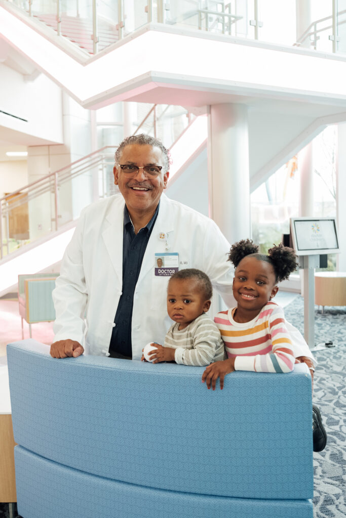 A smiling male doctor in a white lab coat stands behind a blue counter with two happy young children.