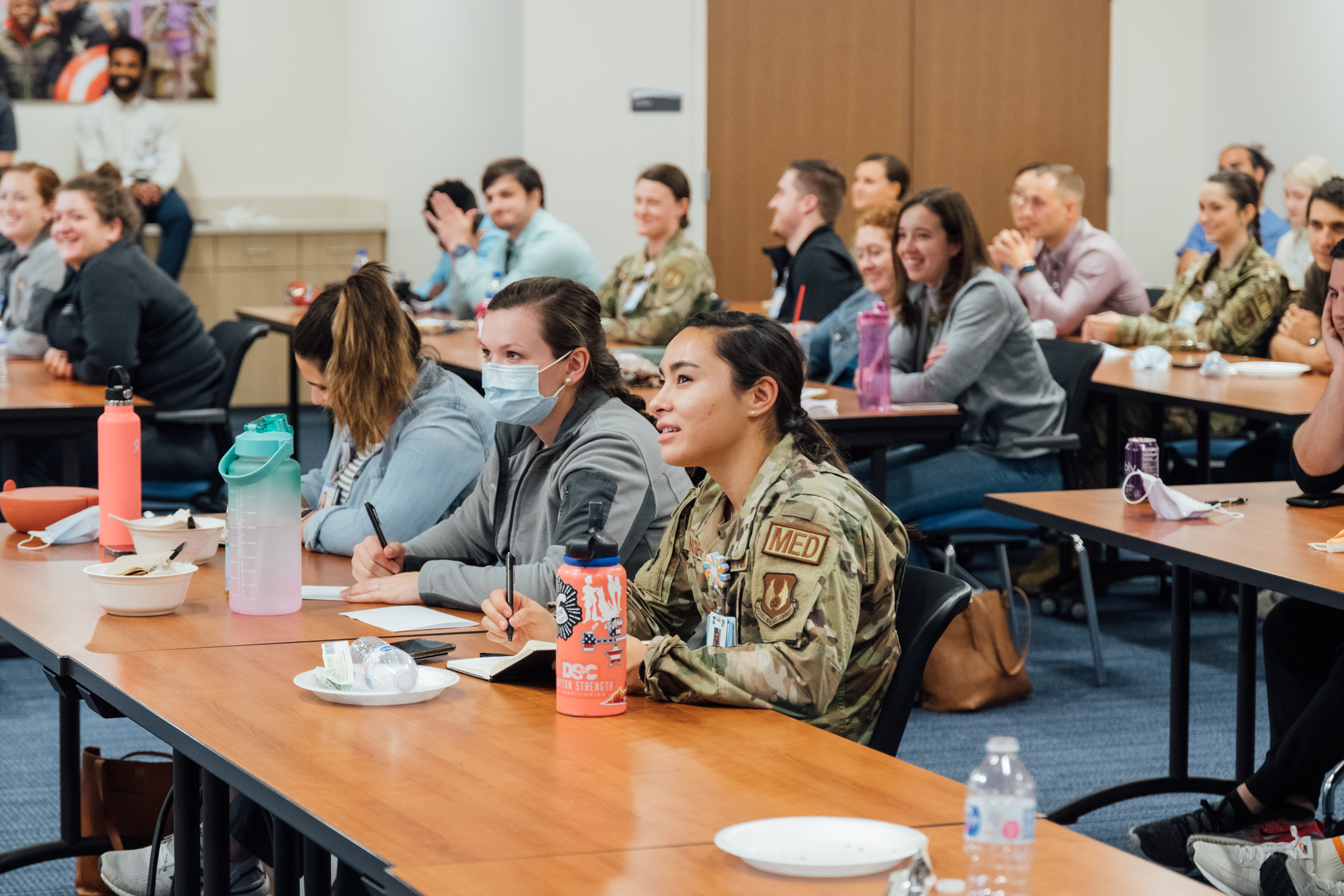 A large group of people, including a woman in a military uniform, sit at tables and smile while attending a lecture or training session.
