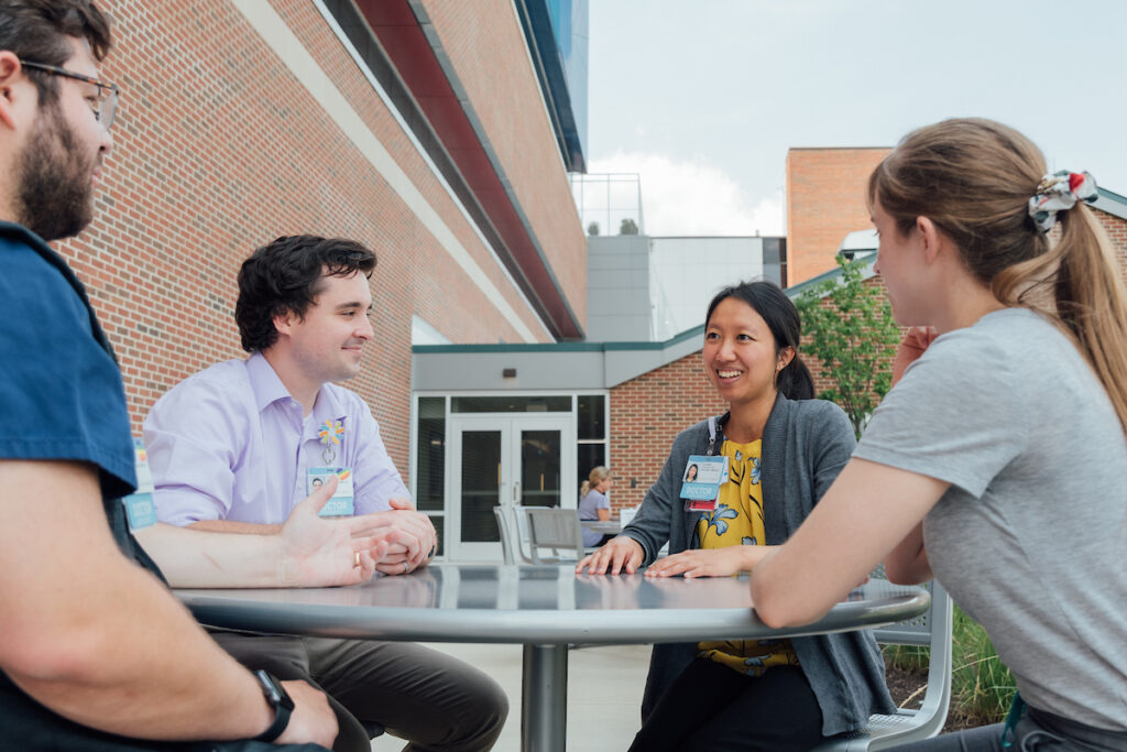 Four medical professionals in casual attire sit around an outdoor table, smiling and talking.