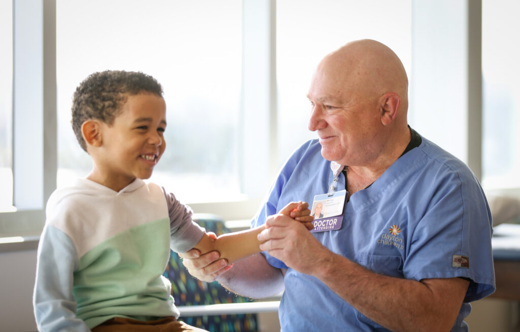 A smiling male doctor in blue scrubs examines the arm of a happy young boy.