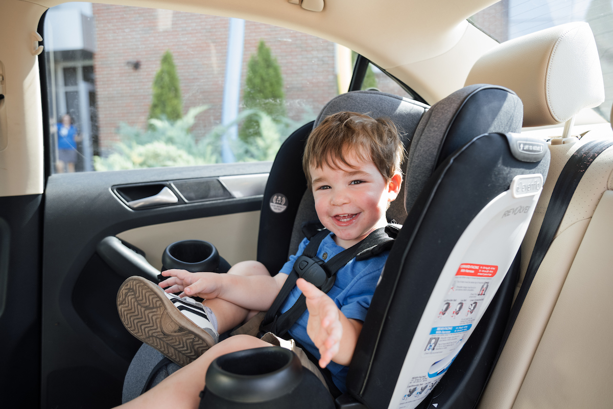 Smiling toddler sitting safely in a car seat inside a vehicle, looking happy and comfortable.

