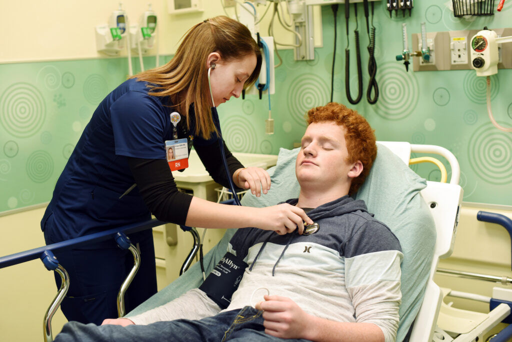 A female nurse with a ponytail uses a stethoscope to examine a male patient with red hair lying in a hospital bed.
