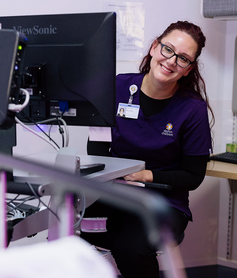 A cheerful female healthcare worker in purple scrubs and glasses smiles while looking over a computer monitor.