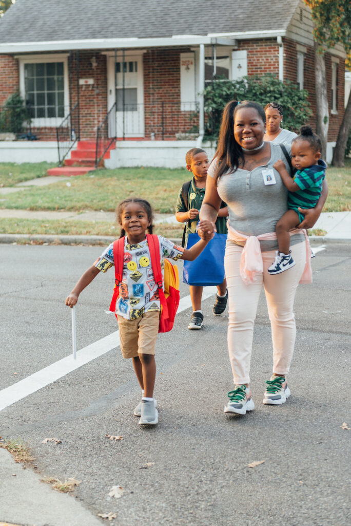 Smiling mother walking with her children to school, holding one child and another carrying a red backpack.

