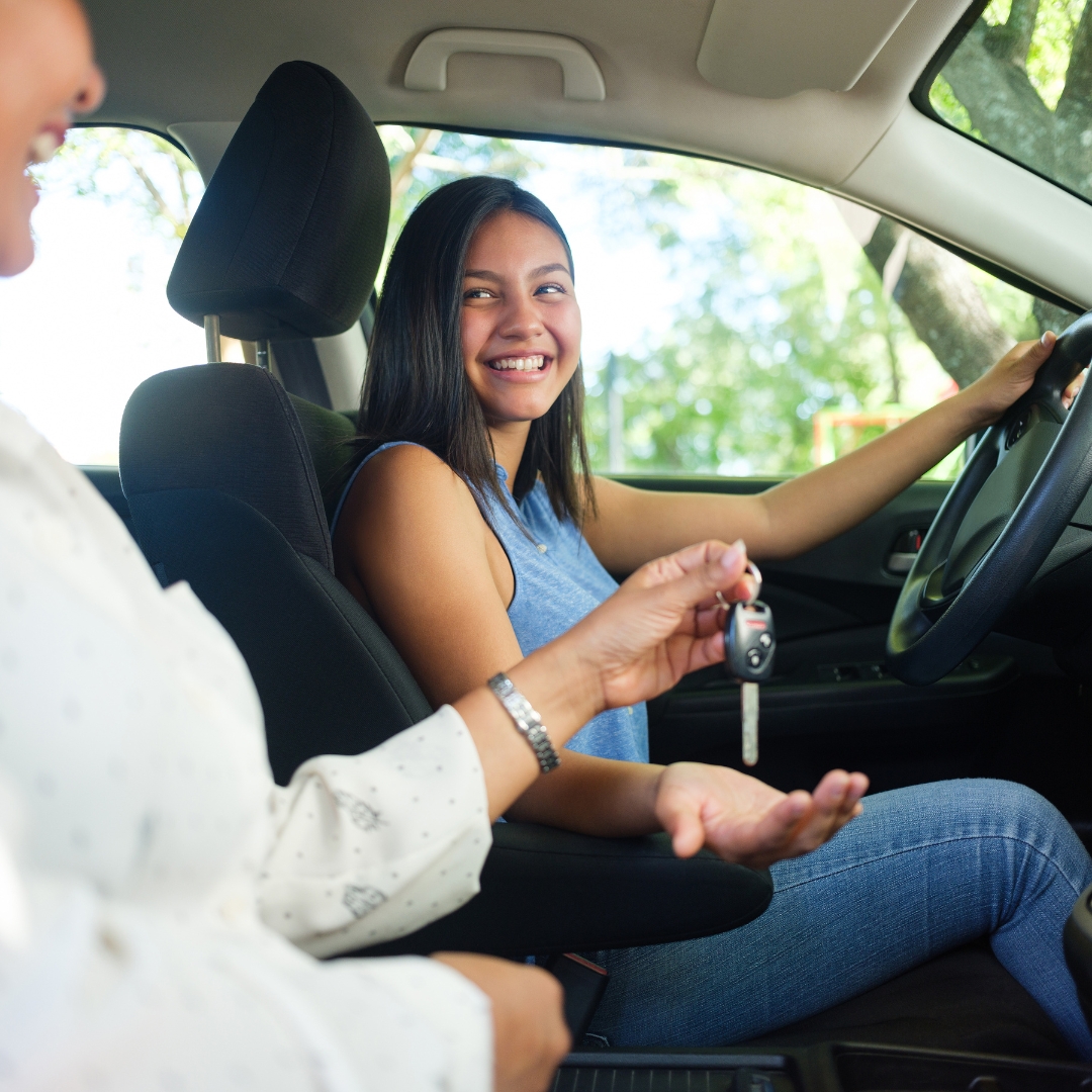Smiling teenage girl in the driver’s seat receiving car keys from her parent, excited to start driving.